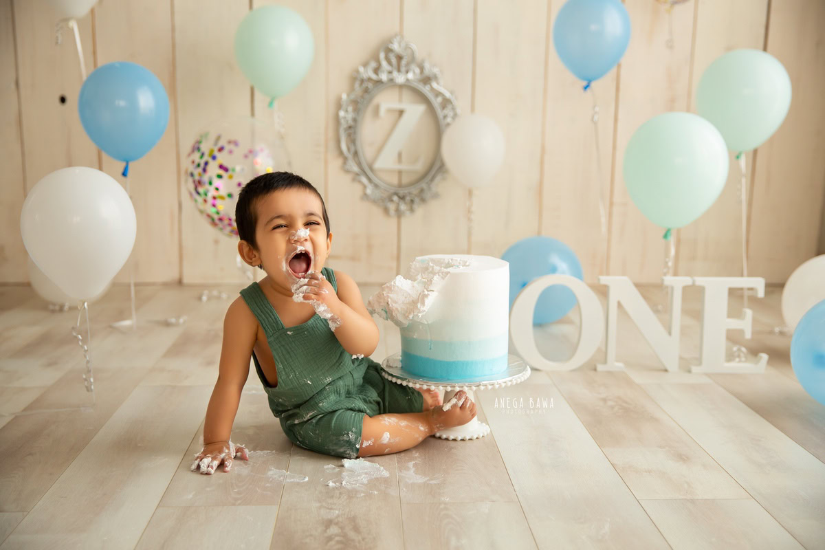 1year old boy enjoying cake smash with a beige backdrop silver frame on the wall and multicolour balloons. Captured during a charming first birthday photo shoot in Delhi Gurgaon by the esteemed family photographer Anega Bawa.