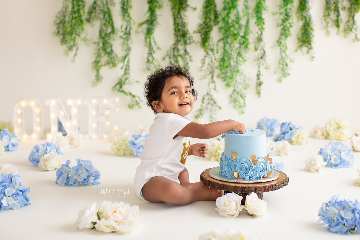 1year old boy enjoying a cake smash with blue and white flowers, leafy fringe on the wall, and a white backdrop. Celebrating his first birthday. Cake Smash photo shoot in Delhi, Gurgaon by Anega Bawa Family Photographer Gurgaon (Delhi NCR).