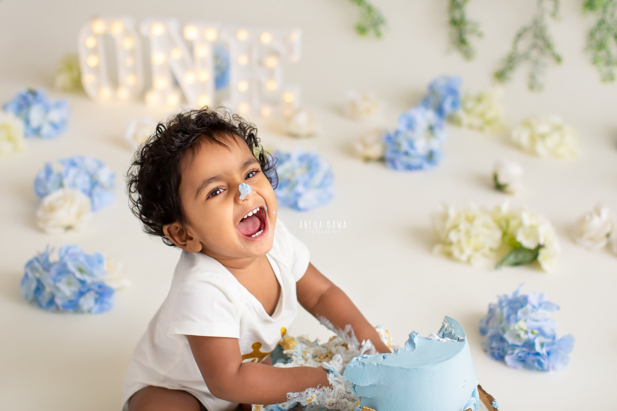 1 year old boy enjoying a cake smash with blue and white flowers against a white backdrop. Captured during a delightful first birthday photography session in Delhi Gurgaon by the renowned family photographer Anega Bawa.