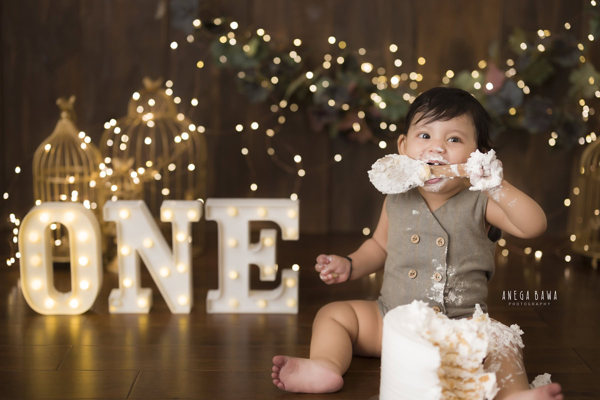 1 year old boy enjoying a cake smash with a brown backdrop and fairy lights. Captured during a charming first birthday photography session in Delhi Gurgaon by the renowned family photographer Anega Bawa.