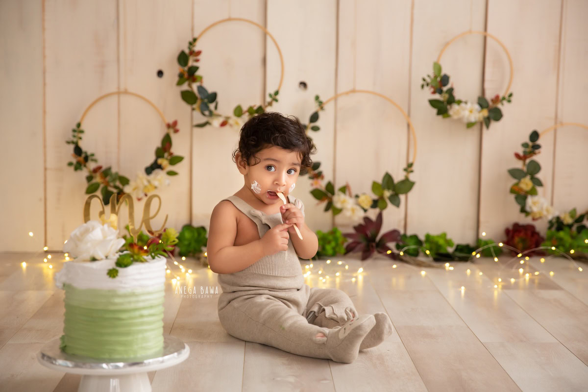 1 year old boy enjoying a cake smash with fairy lights, a beige backdrop, and a wooden floral frame on the wall. Captured during a captivating first birthday photo shoot in Delhi Gurgaon by the renowned family photographer Anega Bawa.