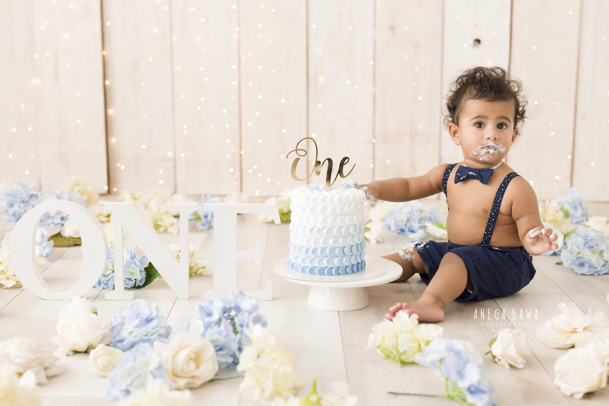 1year old boy with hands in the cake, enjoying a cake smash session with a beige backdrop and blue and white flowers. Celebrating his first birthday. Cake Smash photo shoot in Delhi, Gurgaon by Anega Bawa Family Photographer Gurgaon (Delhi NCR).