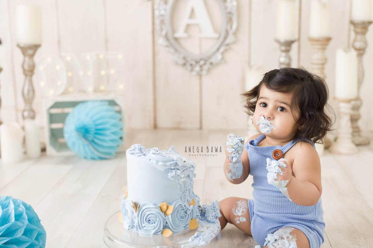 1 year old boy in a blue babysuit with Cake Smash on face, featuring a candle stand, blue pompoms, and a beige backdrop, captured in a charming first birthday photoshoot by Anega Bawa in Delhi, Gurgaon.