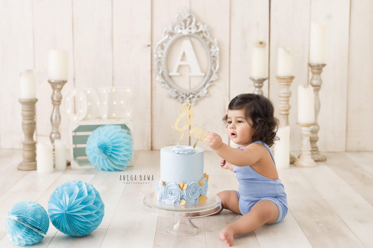 1year old boy in a blue babysuit with a cake smash session, candle stand, blue pompom, and silver alphabet frame on the wall. Celebrating his first birthday. Cake Smash photo shoot in Delhi, Gurgaon by Anega Bawa Family Photographer Gurgaon (Delhi NCR).