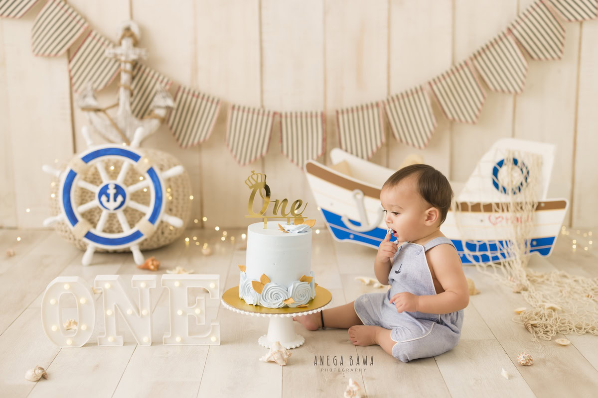 1 year old boy in blue dungarees with Cake Smash, featuring a toy spaceship, fairy lights, and a beige backdrop, captured in delightful first birthday photography by Anega Bawa in Delhi, Gurgaon.