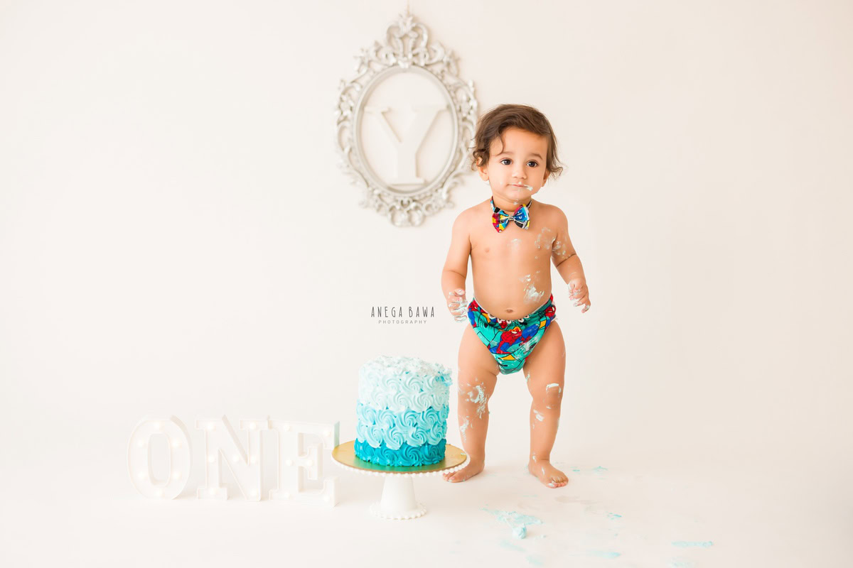 1year old boy in a cute Frenchie and bow, posing with a cake smash session against a white backdrop and an alphabet frame on the wall. Celebrating his first birthday. Cake Smash photo shoot in Delhi, Gurgaon by Anega Bawa Family Photographer Gurgaon (Delhi NCR).