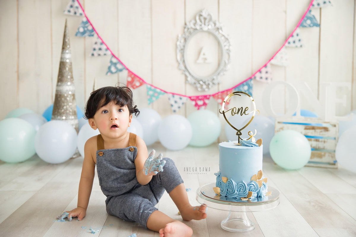 1year old boy in denim dungarees, surrounded by balloons and a cake smash setup, with a beige backdrop and a silver alphabet frame on the wall. Celebrating his first birthday in style. Cake Smash photo shoot in Delhi, Gurgaon by Anega Bawa Family Photographer Gurgaon (Delhi NCR).
