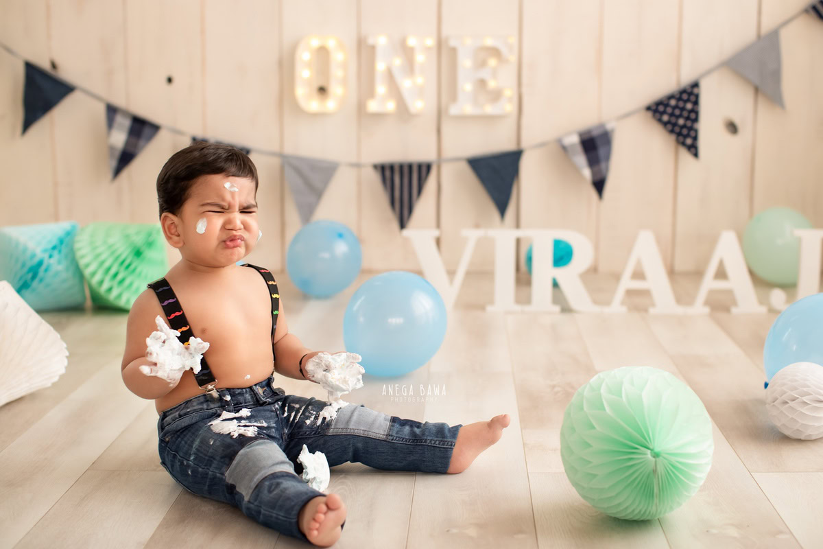 1 year old boy with cake smash on his face and hands, name frame on the floor, blue and green pompom decorations, and a beige backdrop. Captured during a charming first birthday photography session in Delhi Gurgaon by the renowned family photographer Anega Bawa.