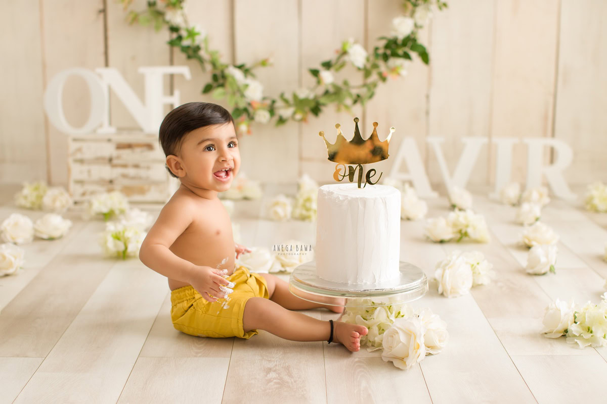 1 year old boy with name frame on floor white flowers Cake Smash floral curve fringe on wall beige backdrop first birthday photography in Delhi Gurgaon. Captured by Anega Bawa family photographer Gurgaon (Delhi NCR)