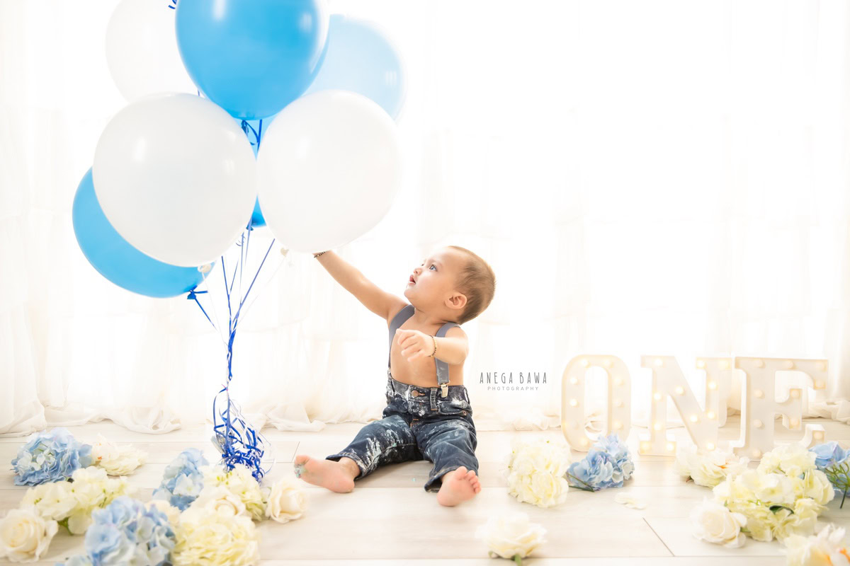 1 year old boy playing with balloons Cake Smash on hands white backdrop flowers on floor first birthday photoshoot in Delhi Gurgaon. Captured by Anega Bawa family photographer Gurgaon (Delhi NCR)