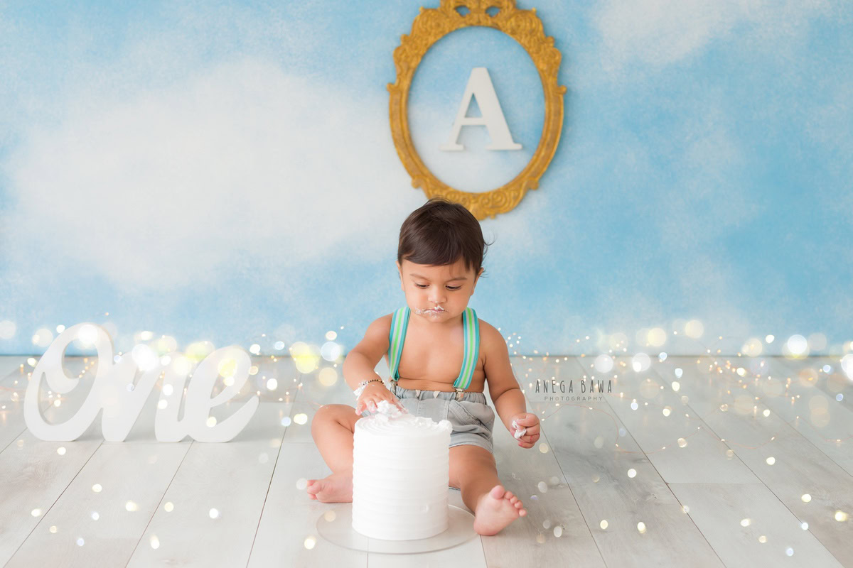 1 year boy playing with a cake smash, set against a blue and white backdrop with an alphabet frame on the wall and fairy lights during a first birthday photography session by well-known Anega Bawa Photography in Delhi, Gurgaon.