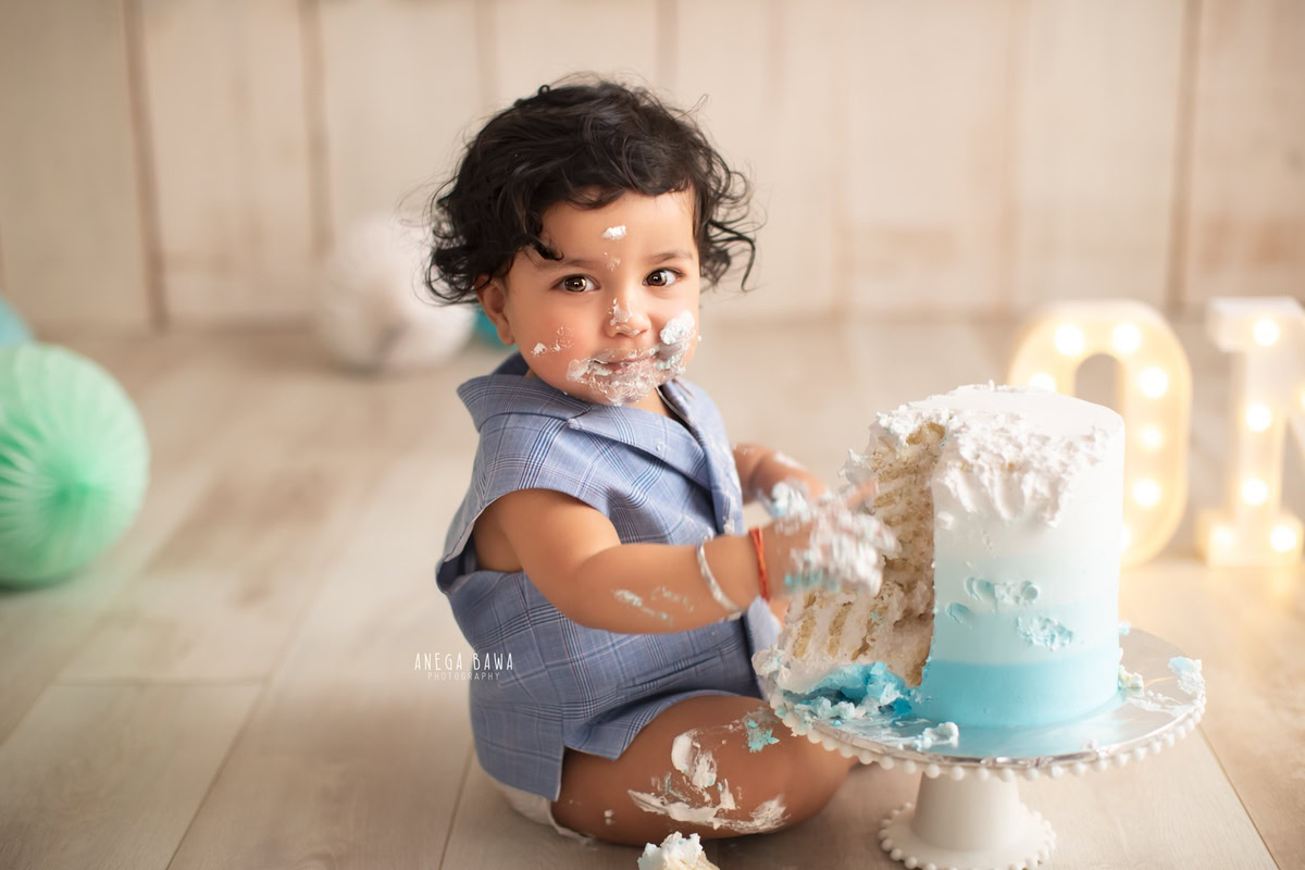 1 year old boy playing with a cake smash setup, featuring a beige backdrop and blue and green pompoms. Celebrating his first birthday. Cake Smash photo shoot in Delhi, Gurgaon by Anega Bawa Family Photographer Gurgaon (Delhi NCR).
