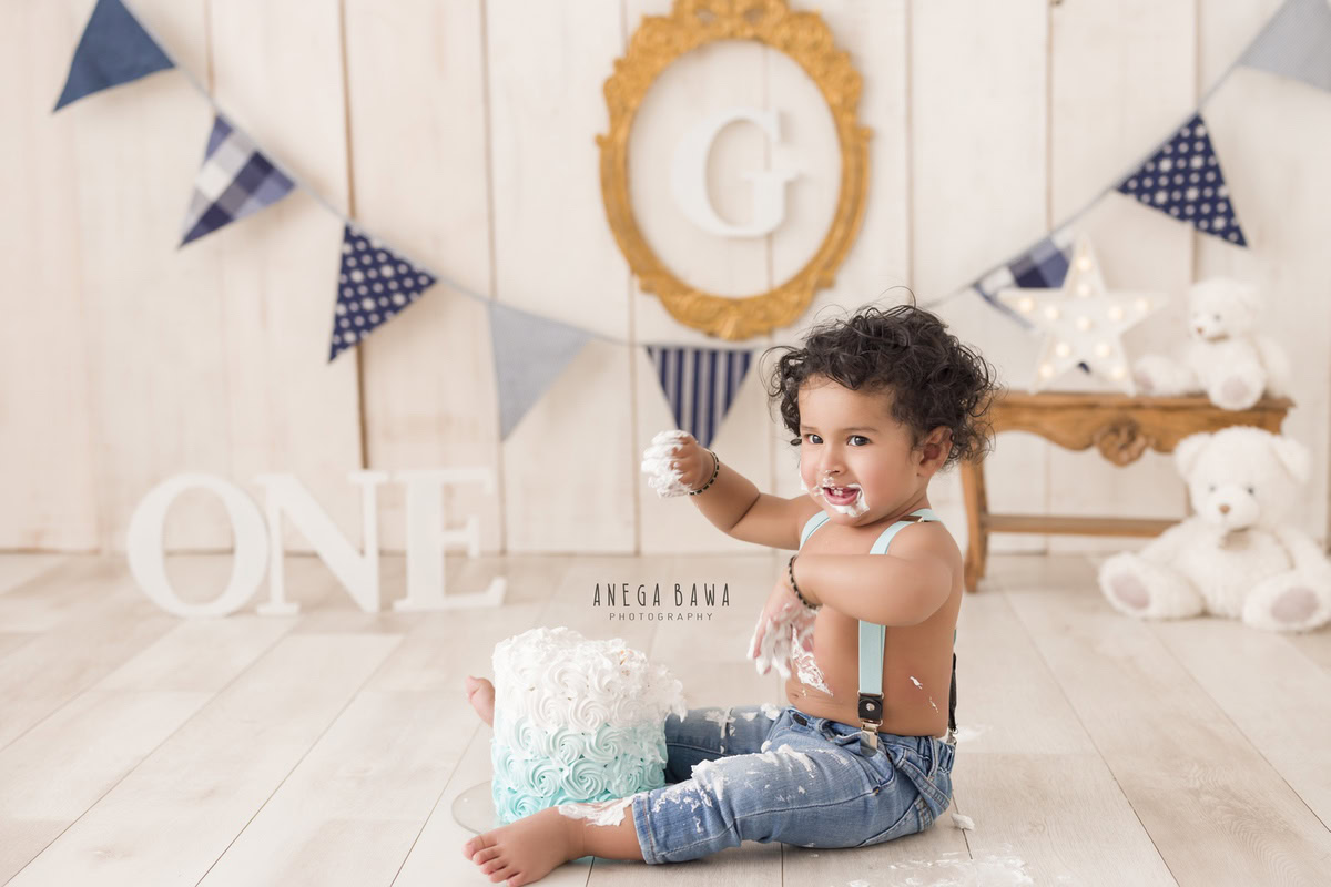 1 year old boy playing with Cake Smash, set against a beige backdrop with a teddybear and an alphabet frame on the wall, captured in a delightful first birthday photoshoot by Anega Bawa in Delhi, Gurgaon.