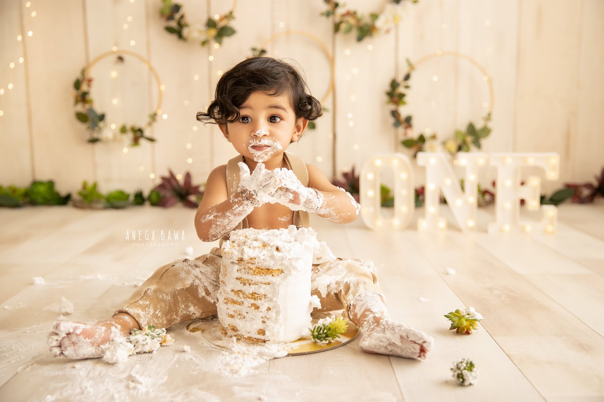 1 year old boy playing with Cake Smash, beige backdrop, wooden leafy frame on wall, first birthday photography in Delhi, Gurgaon. Captured by Anega Bawa family photographer Gurgaon (Delhi NCR)
