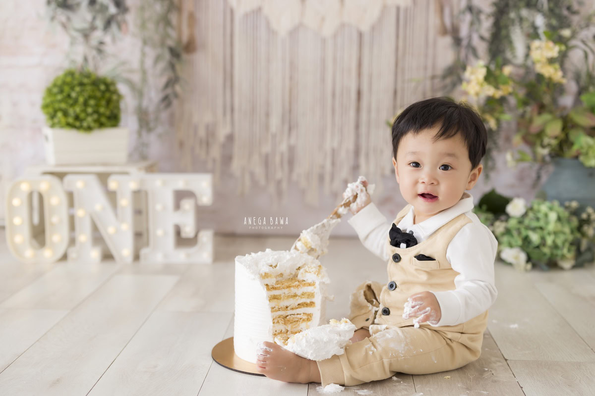 1 year boy playing with a cake smash against a beige net backdrop during a first birthday photoshoot by well-known Anega Bawa Photography in Delhi, Gurgaon.
