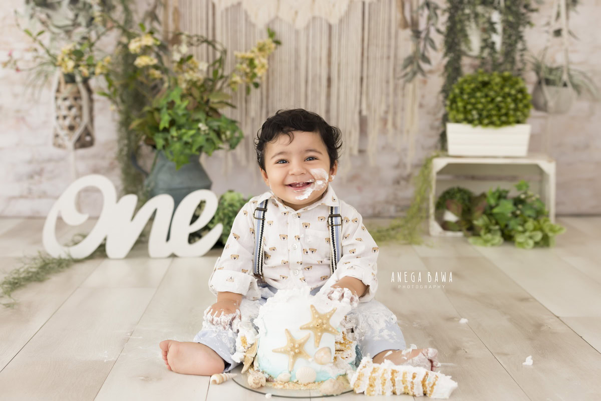 1 year old boy playing with cake smash against a beige net backdrop, green vase, and leafy fringes. Captured during an engaging first birthday photography session in Delhi Gurgaon by the renowned family photographer Anega Bawa.
