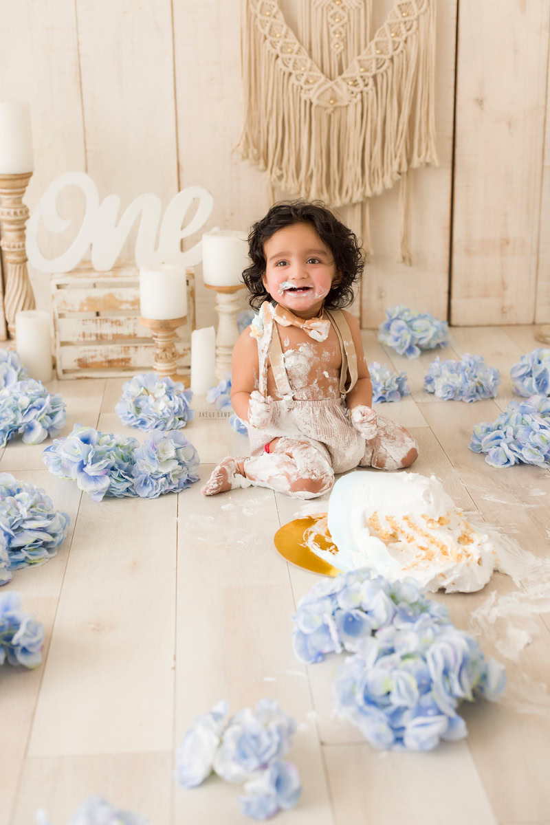 1 year boy playing with a cake smash, surrounded by blue flowers on the floor and a candle stand against a beige backdrop during a first birthday photoshoot by well-known Anega Bawa Photography in Delhi, Gurgaon.
