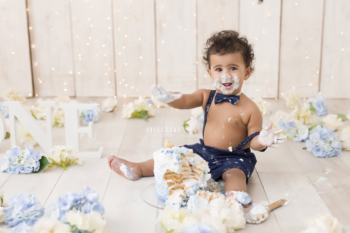 1 year old boy playing with Cake Smash, blue and white flowers, beige backdrop, fairy lights, first birthday photoshoot in Delhi, Gurgaon. Captured by Anega Bawa family photographer Gurgaon (Delhi NCR)
