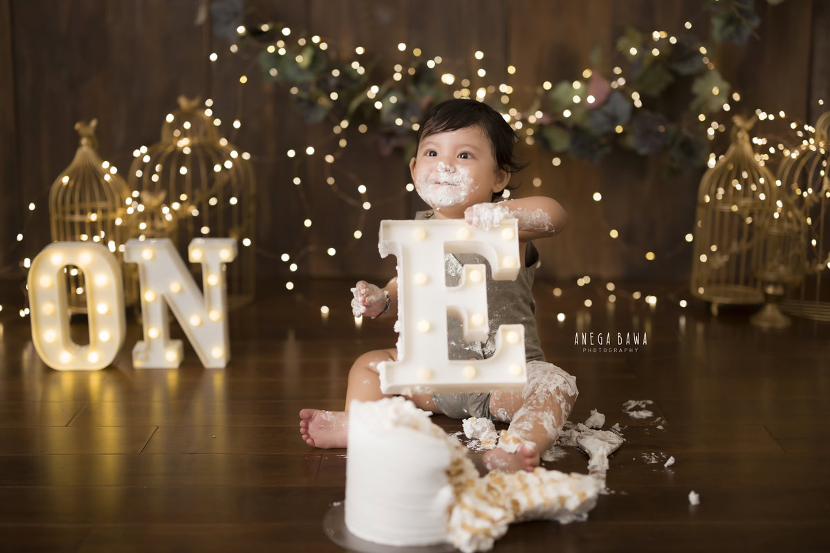 1 year old boy playing with Cake Smash, set against a brown backdrop with golden lights, captured in captivating first birthday photography by Anega Bawa in Delhi, Gurgaon.