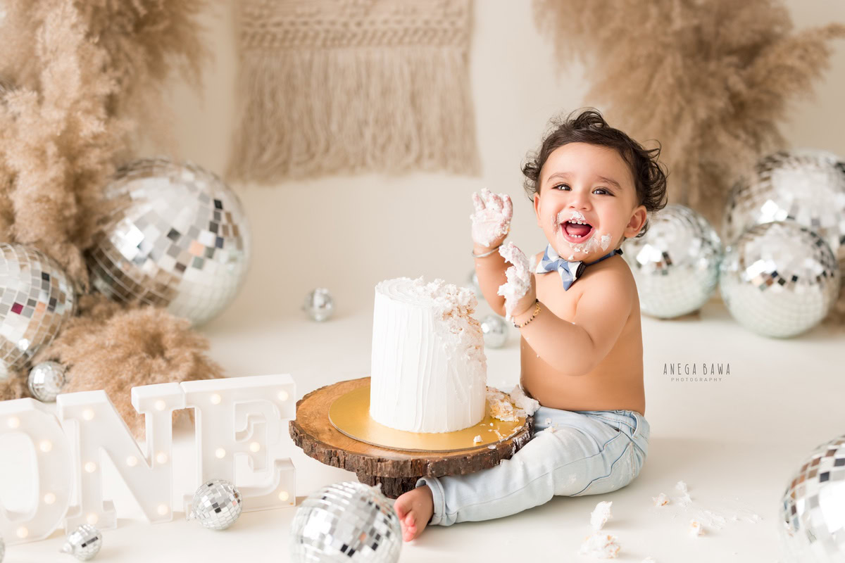1 year old boy playing with cake smash, surrounded by bushes and discoballs, against a beige backdrop. Captured during a vibrant first birthday photography session in Delhi Gurgaon by the renowned family photographer Anega Bawa.