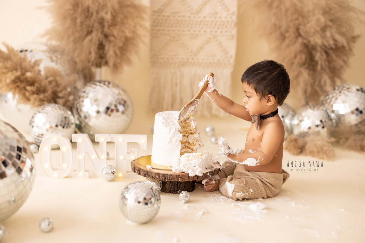 1 year old boy playing with Cake Smash, bushes, disco balls, beige backdrop, first birthday photoshoot in Delhi, Gurgaon. Captured by Anega Bawa family photographer Gurgaon (Delhi NCR)