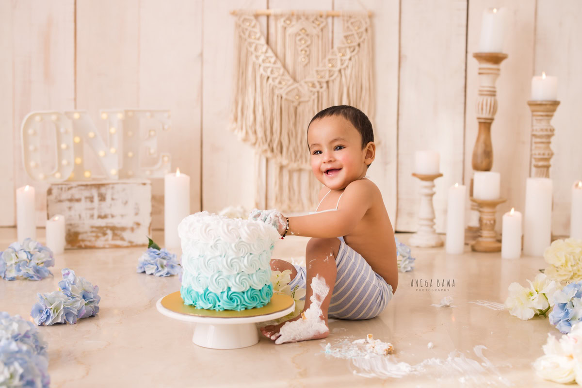 1 year old boy playing with Cake Smash, featuring a candle stand with white candles, a beige backdrop, and blue flowers on the floor, captured in a delightful first birthday photoshoot by Anega Bawa in Delhi, Gurgaon.