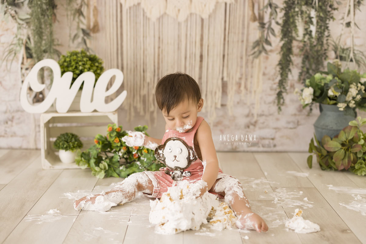 1 year old boy playing with cake smash, against a net beige backdrop, green vase, and leafy fringes. Captured during a delightful first birthday photo shoot in Delhi Gurgaon by the renowned family photographer Anega Bawa.