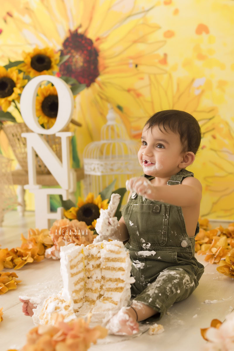 1 year old boy playing with cake smash, set against a sunflower backdrop. Captured during a joyful first birthday photography session in Delhi Gurgaon by the renowned family photographer Anega Bawa.