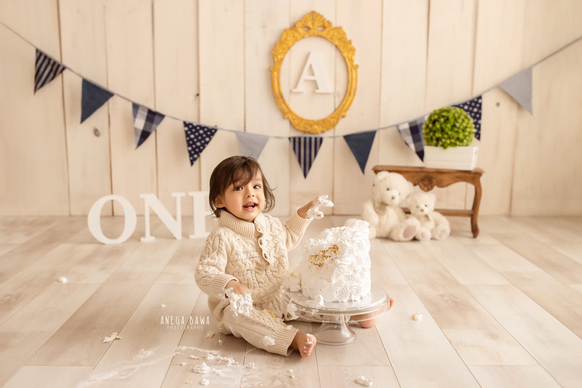 1 year old boy playing with a cake smash setup, featuring teddy bears, a wooden stool, and a beige backdrop. Celebrating his first birthday. Cake Smash photo shoot in Delhi, Gurgaon by Anega Bawa Family Photographer Gurgaon (Delhi NCR).