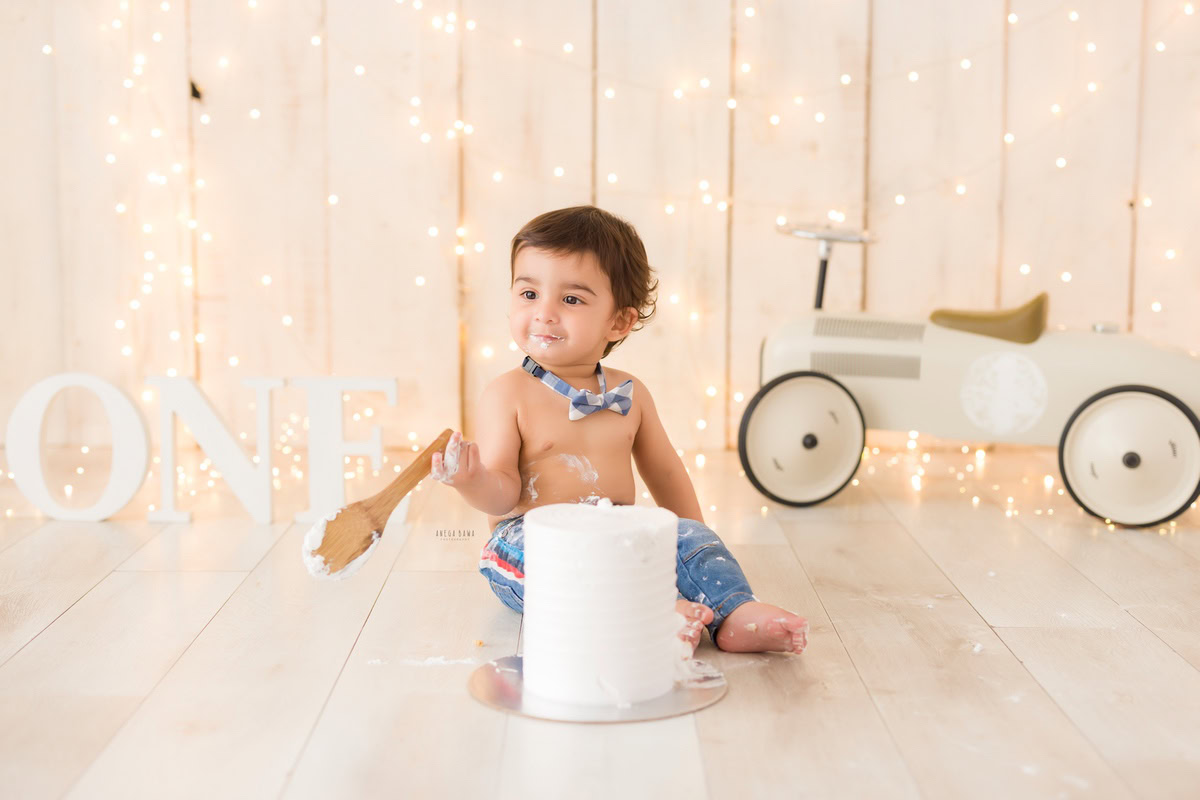 1 year old boy playing with Cake Smash, featuring a toy scooter and fairy lights against a beige backdrop, captured in enchanting first birthday photography by Anega Bawa in Delhi, Gurgaon.