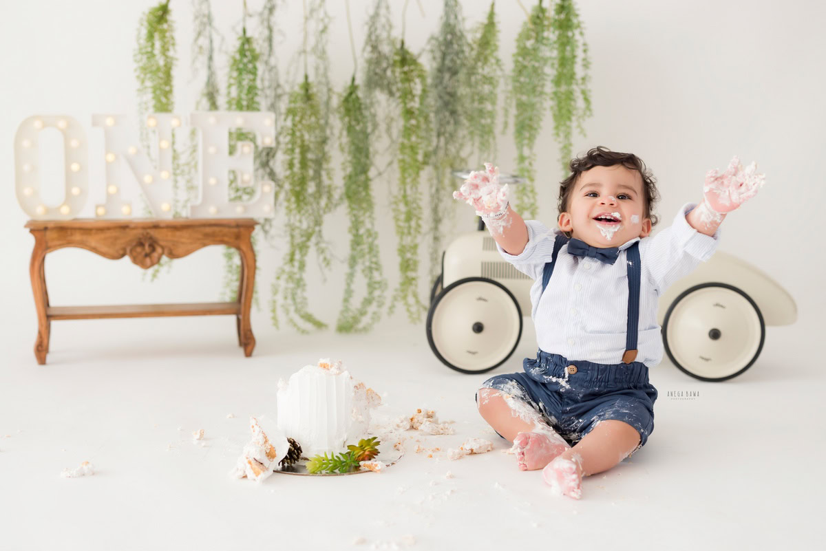 1year old boy playing with a cake smash toy scooter against a white backdrop with leafy fringes, captured by Anega Bawa during a Cake Smash photoshoot in Delhi, Gurgaon