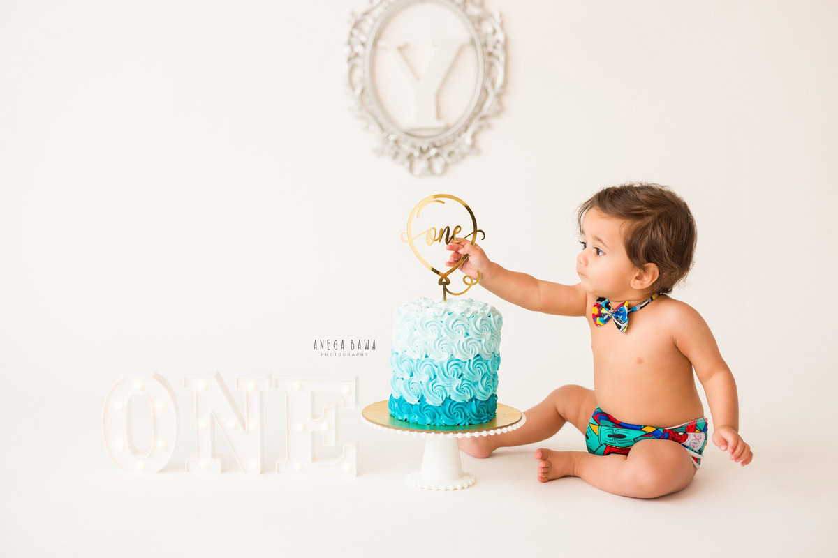 1year old boy playing with a cake smash setup against a white backdrop featuring a silver alphabet frame on the wall and a cute bow, captured by Anega Bawa during a Cake Smash photoshoot in Delhi, Gurgaon