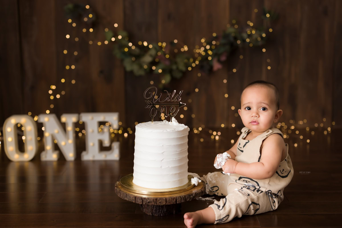1year old boy posing with a cake smash setup against a brown backdrop with fairy lights, captured by Anega Bawa during a Cake Smash photoshoot in Delhi, Gurgaon