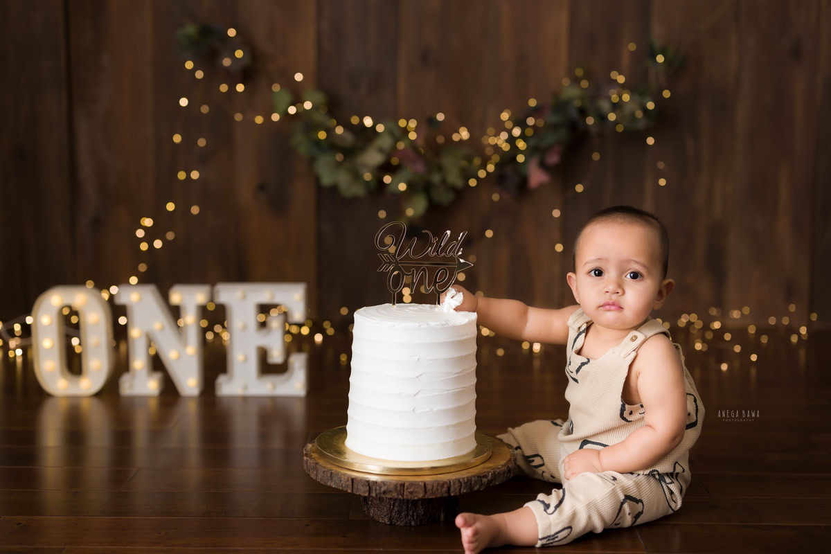 1year old boy posing with a cake smash setup against a brown backdrop with fairy lights, captured by Anega Bawa during a Cake Smash photoshoot in Delhi, Gurgaon