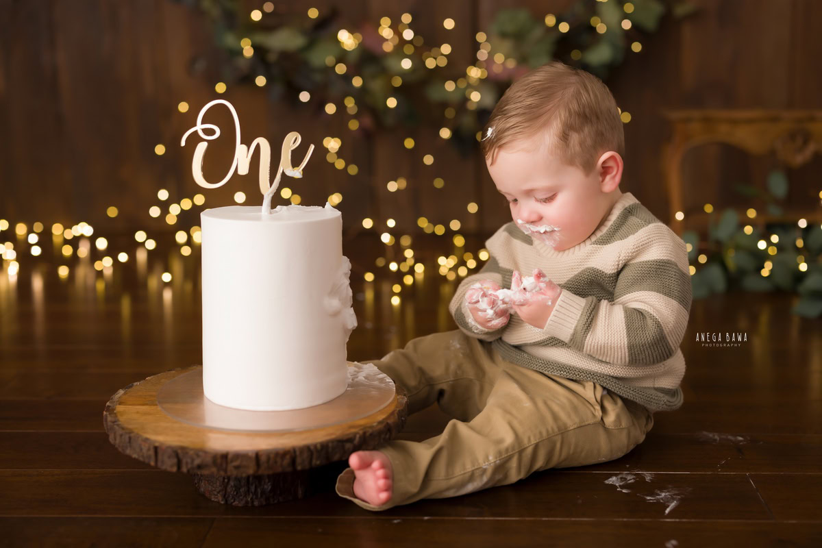 1 year boy posing with a cake smash, set against a brown backdrop with fairy lights during a first birthday photoshoot by well-known Anega Bawa Photography in Delhi, Gurgaon.