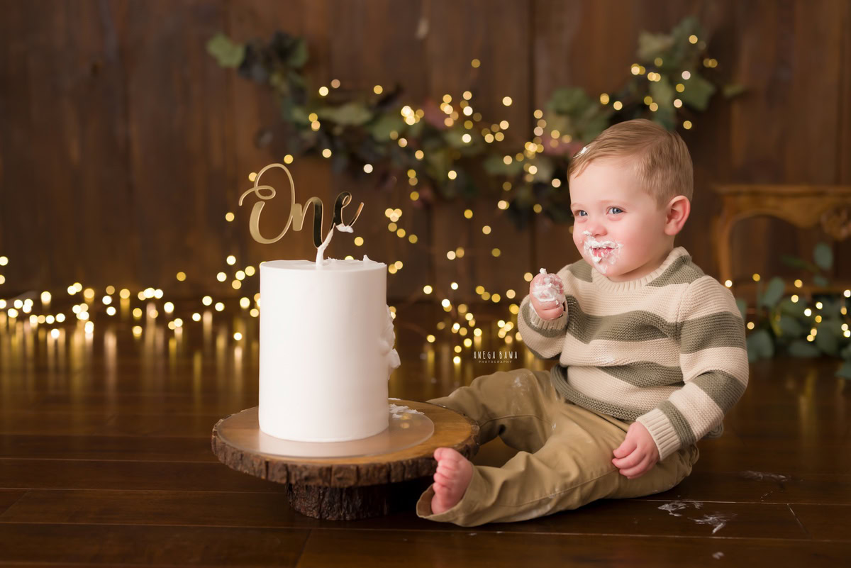 1year old boy posing with a cake smash setup against a brown backdrop with fairy lights, captured by Anega Bawa during a Cake Smash photoshoot in Delhi, Gurgaon