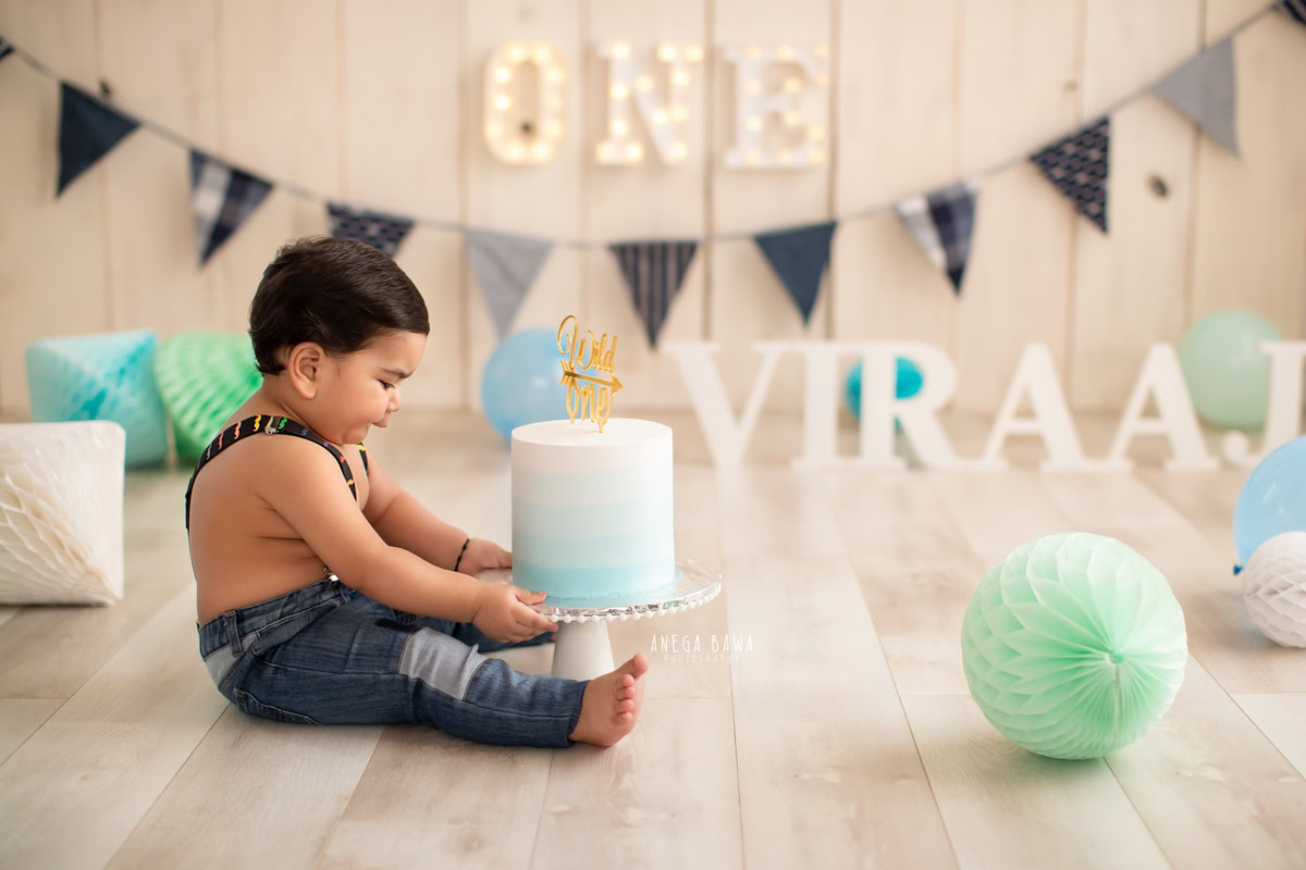 1 year old boy posing with a cake smash setup, featuring green and blue pompoms against a beige backdrop. Celebrating his first birthday. Cake Smash photo shoot in Delhi, Gurgaon by Anega Bawa Family Photographer Gurgaon (Delhi NCR).