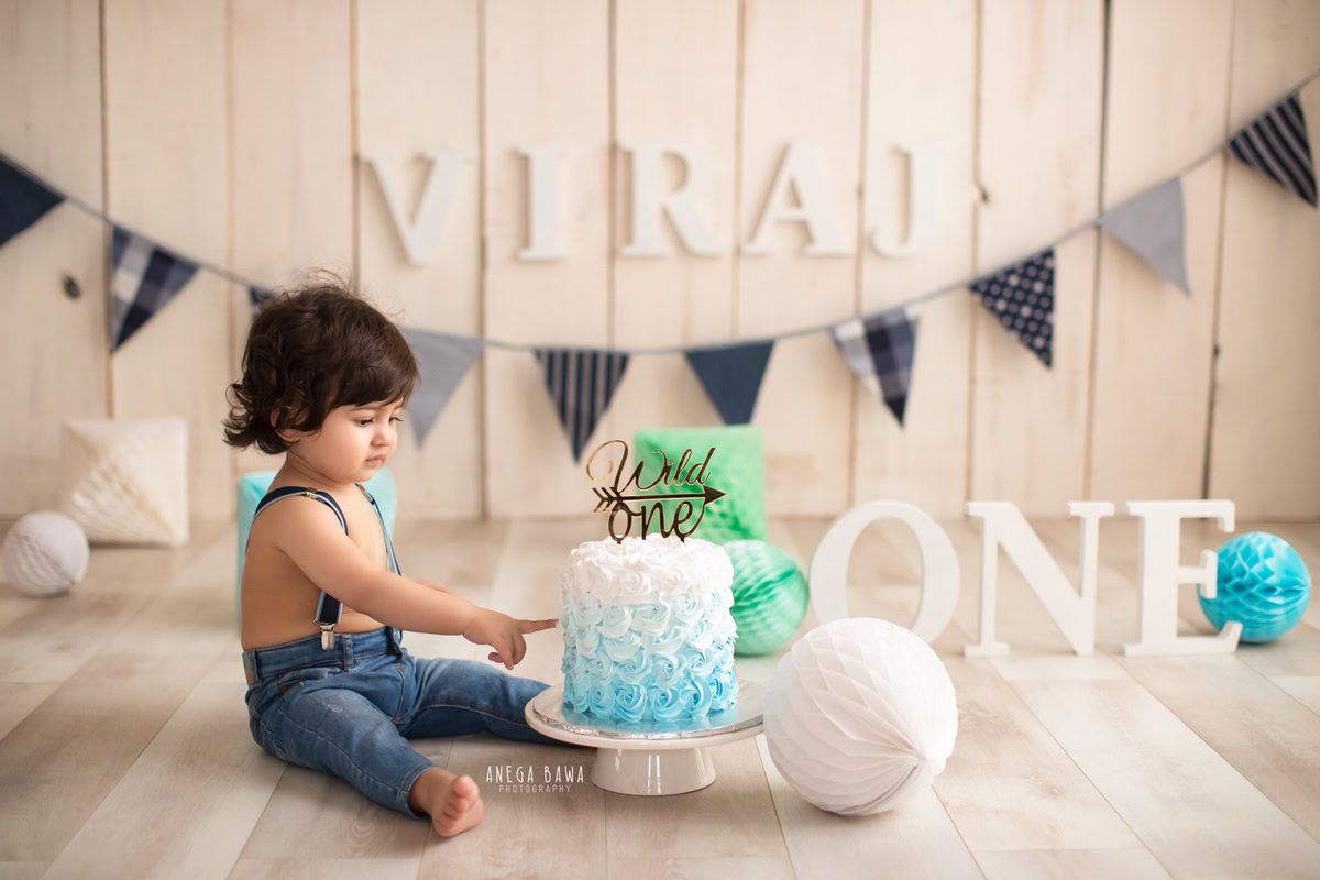 1 year old boy posing with a cake smash setup, featuring pompoms and a name frame on the wall. Celebrating his first birthday. Cake Smash photo shoot in Delhi, Gurgaon by Anega Bawa Family Photographer Gurgaon (Delhi NCR).