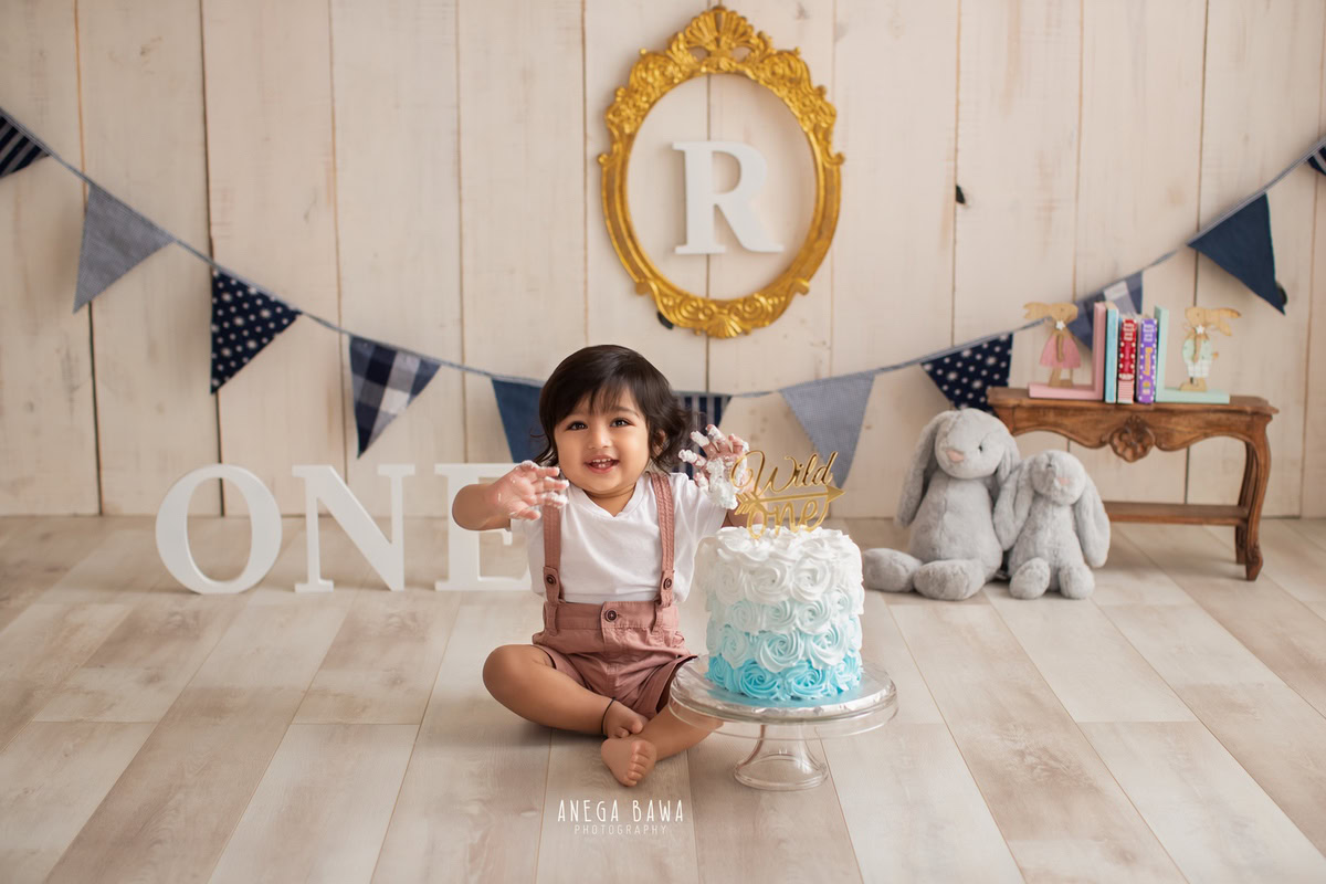 1 year old boy posing with a cake smash setup, featuring teddy bears, a beige backdrop, and an alphabet frame on the wall. Celebrating his first birthday. Cake Smash photo shoot in Delhi, Gurgaon by Anega Bawa Family Photographer Gurgaon (Delhi NCR).