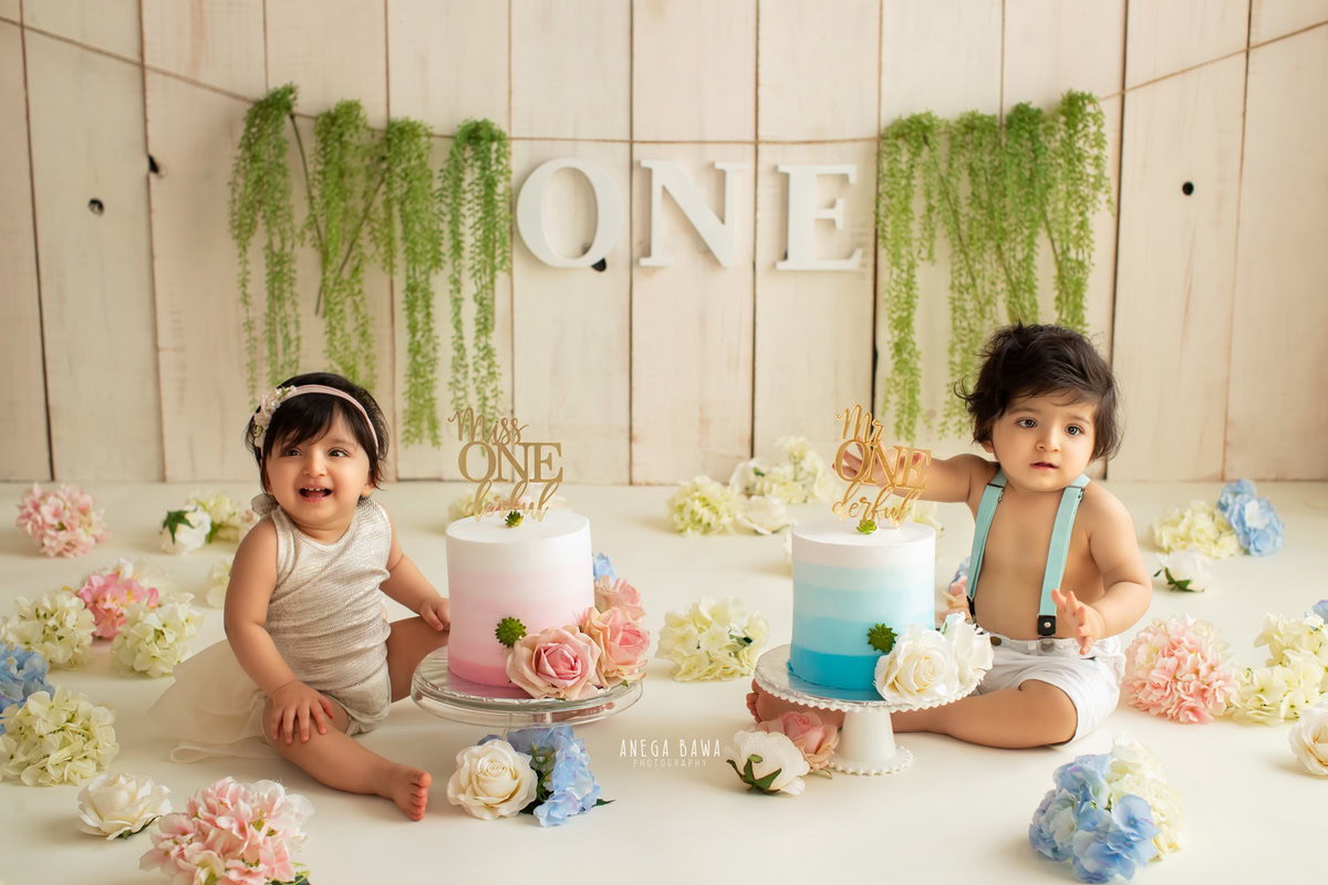 1 year old boy and sibling with two cake smash setups, featuring a leafy fringe on the wall, flowers on the floor, and a beige backdrop. Celebrating their first birthday. Cake Smash photo shoot in Delhi, Gurgaon by Anega Bawa Family Photographer Gurgaon (Delhi NCR).