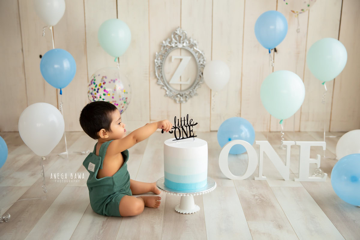 1 year old boy sitting on the floor with a cake smash setup, featuring balloons, a beige backdrop, and a silver frame on the wall. Celebrating his first birthday. Cake Smash photo shoot in Delhi, Gurgaon by Anega Bawa Family Photographer Gurgaon (Delhi NCR).
