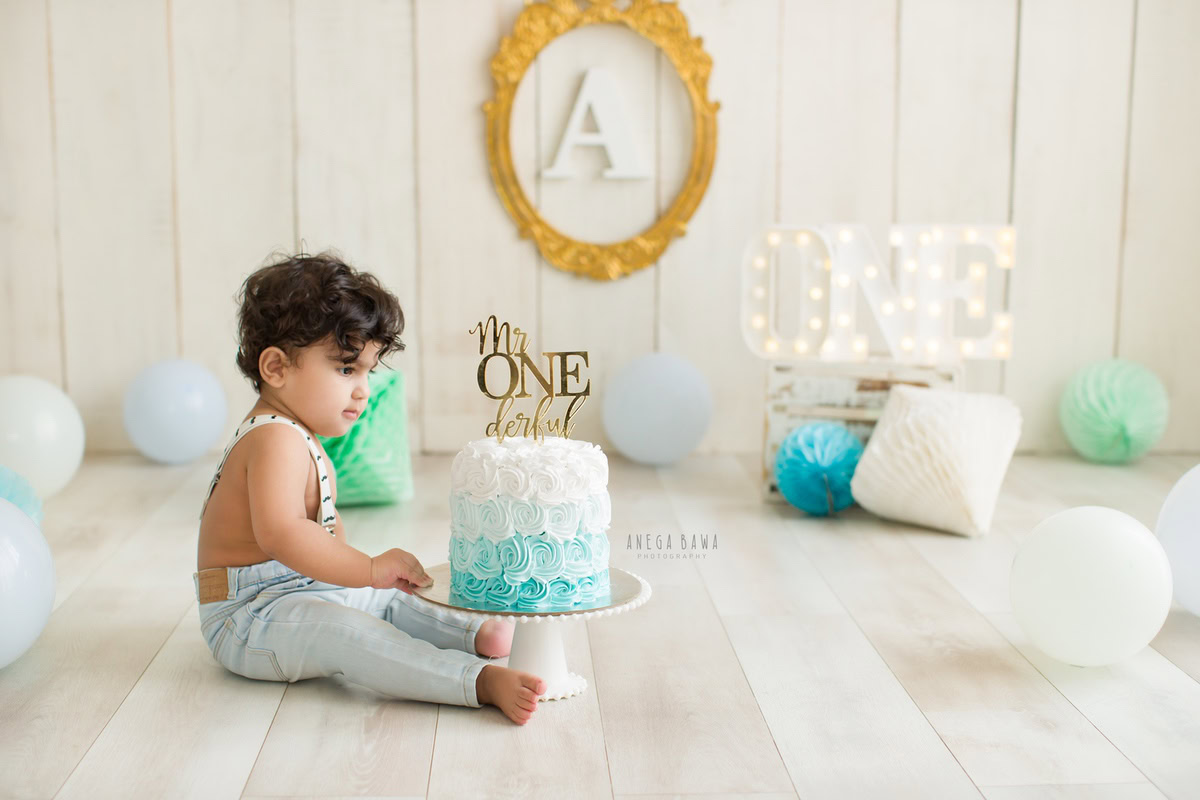 1 year old boy sitting on the floor with a cake smash setup, featuring an alphabet frame on the wall, blue and white pompoms, and a beige backdrop. Celebrating his first birthday. Cake Smash photo shoot in Delhi, Gurgaon by Anega Bawa Family Photographer Gurgaon (Delhi NCR).