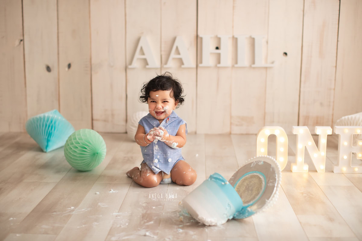 1 year old boy sitting on floor with Cake Smash, blue and green pompom, name frame on wall, beige backdrop, first birthday photography in Delhi, Gurgaon. Captured by Anega Bawa family photographer Gurgaon (Delhi NCR)