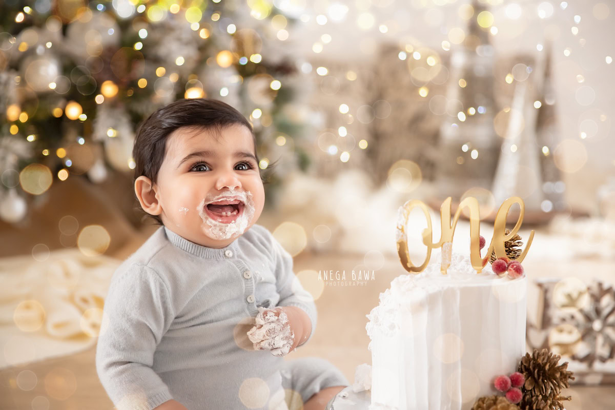 1 year old boy sitting with a cake smash setup, wearing a grey babysuit, surrounded by fairy lights and a Christmas-themed backdrop. Celebrating his first birthday. Cake Smash photo shoot in Delhi, Gurgaon by Anega Bawa Family Photographer Gurgaon (Delhi NCR).