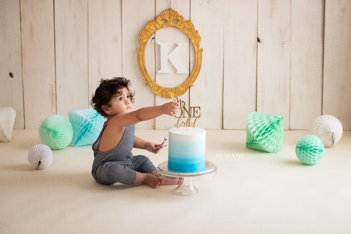 1 year old boy sitting with a cake smash setup, featuring a golden frame on the wall, pompoms, and a beige backdrop. Celebrating his first birthday. Cake Smash photo shoot in Delhi, Gurgaon by Anega Bawa Family Photographer Gurgaon (Delhi NCR).