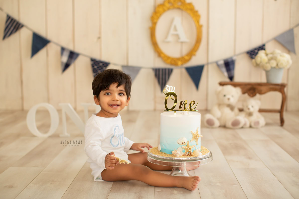 1 year old boy sitting with a cake smash setup, featuring teddy bears, an alphabet frame on the wall, and a beige backdrop. Celebrating his first birthday. Cake Smash photo shoot in Delhi, Gurgaon by Anega Bawa Family Photographer Gurgaon (Delhi NCR).