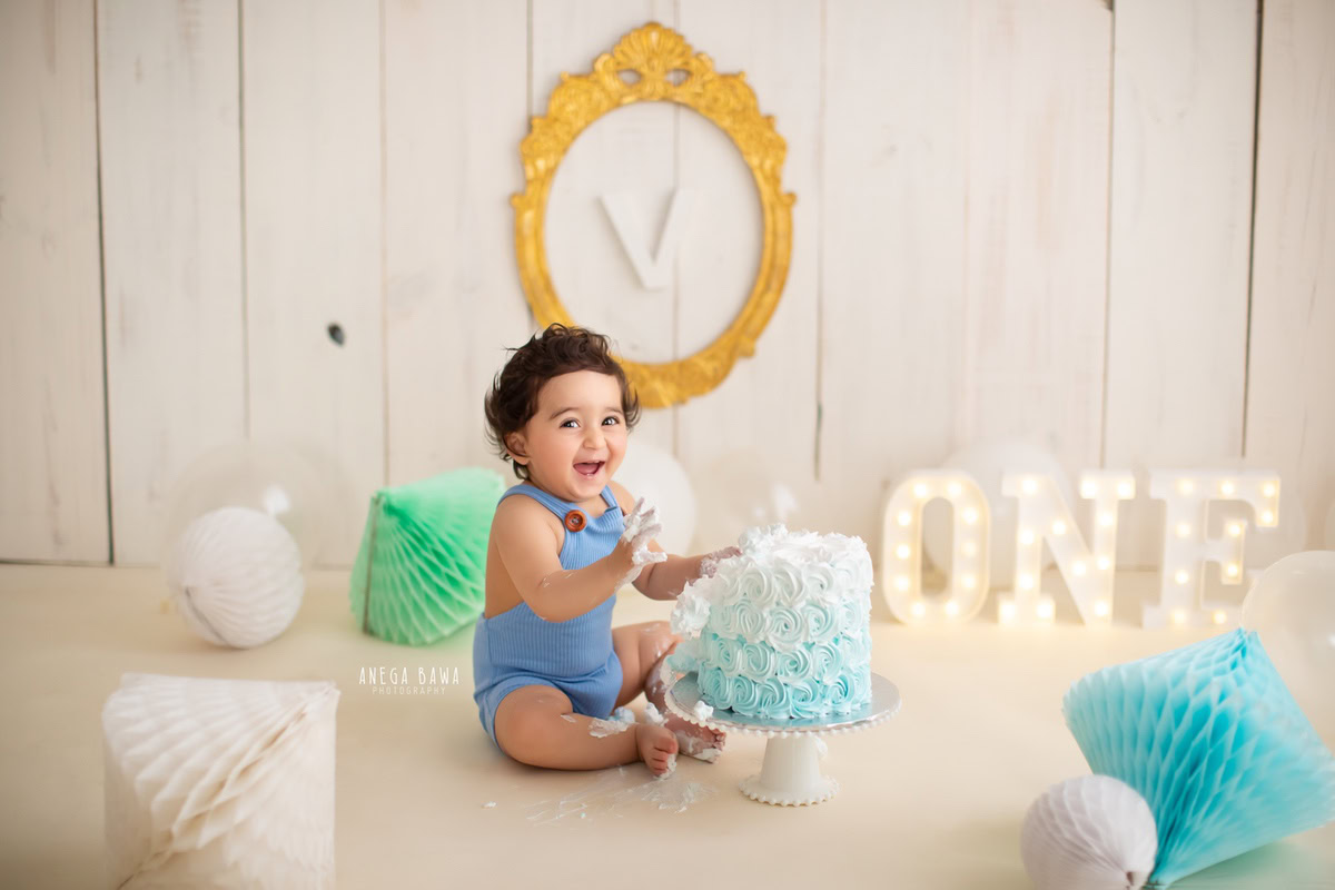 1 year old boy smiling with a cake smash setup, featuring pompoms, a golden alphabet frame on the wall, and a beige backdrop. Celebrating his first birthday. Cake Smash photo shoot in Delhi, Gurgaon by Anega Bawa Family Photographer Gurgaon (Delhi NCR).
