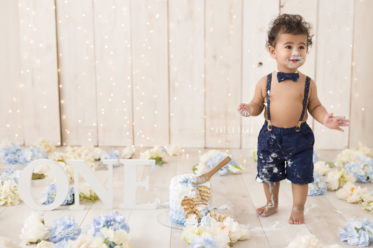 1year old boy standing with a cake smash setup against a beige backdrop with fairy lights and blue and white flowers, captured by Anega Bawa during a Cake Smash photoshoot in Delhi, Gurgaon