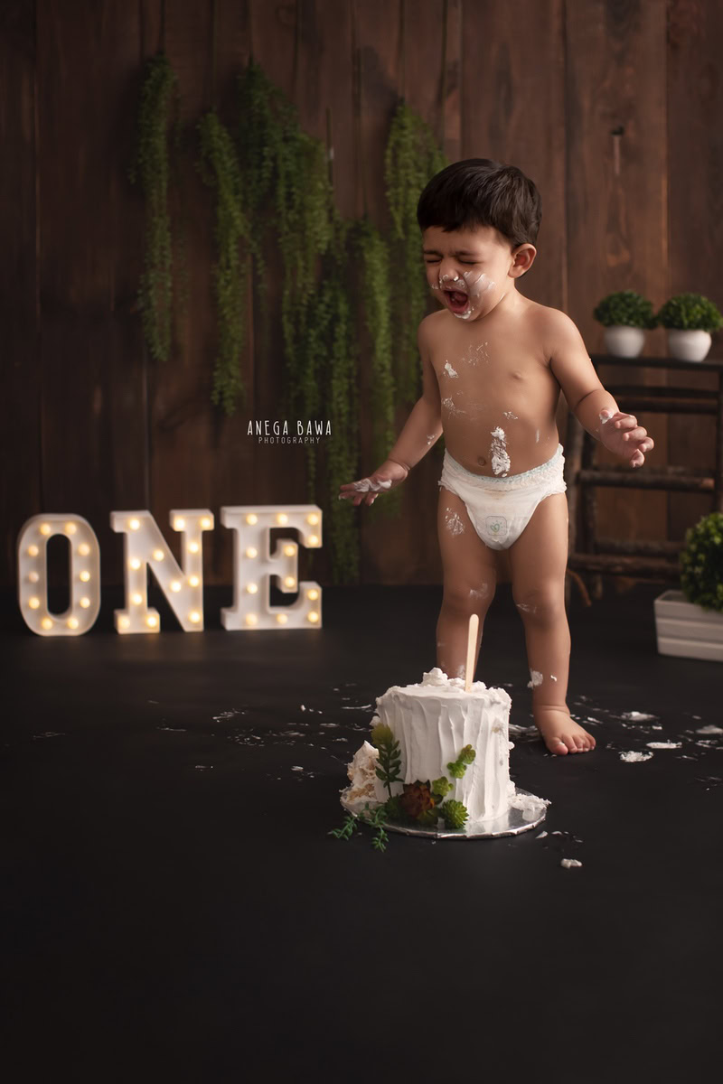 year old boy standing with Cake Smash, brown backdrop, first birthday photography in Delhi, Gurgaon. Captured by Anega Bawa family photographer Gurgaon (Delhi NCR)