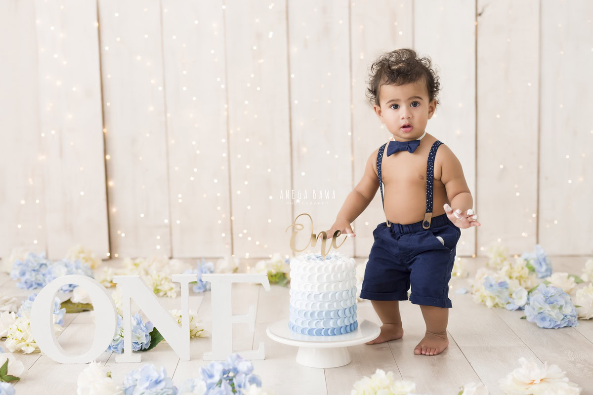 1 year old boy standing with colorful Cake Smash, vibrant blue and white flowers, elegant beige backdrop, twinkling fairy lights, first birthday photoshoot in Delhi, Gurgaon. Captured by Anega Bawa family photographer Gurgaon (Delhi NCR)